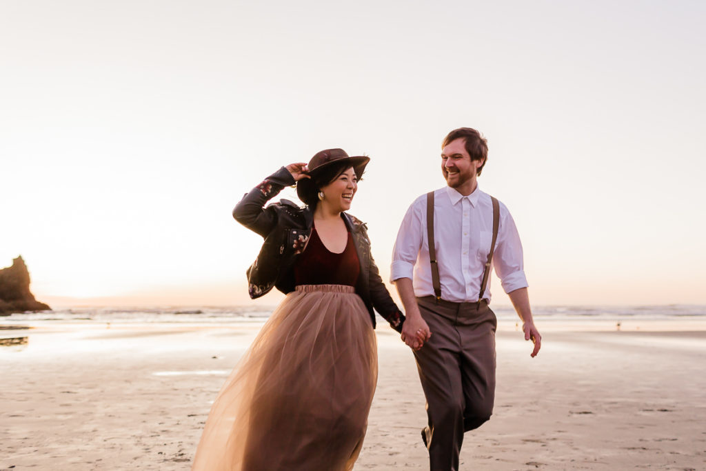 bride in a hat and groom running together at cannon beach