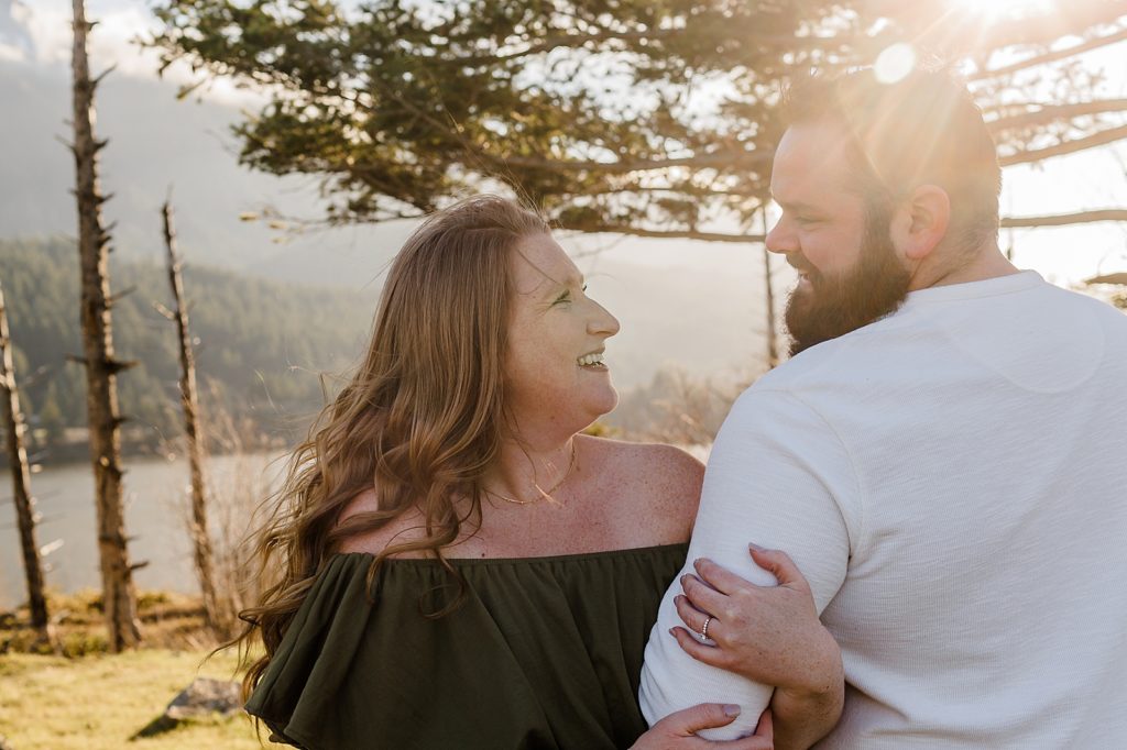 Couple holding each others arms facing each other and looking into each others eyes on the mountains with lens flare Government Cove Oregon Engagement Photography captured by Oregon Engagement Photographer Simply Wandering Photography 