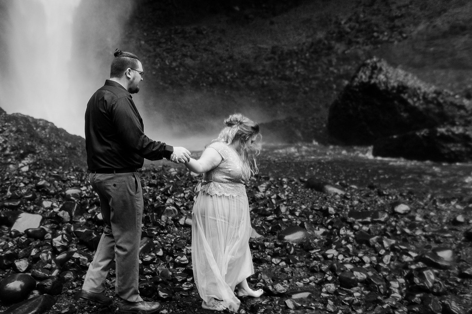 B&W of woman holding man's hand and leading him through rocky waterfall 