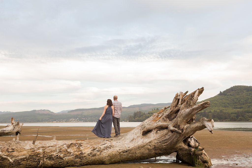 Couple standing on fallen dead tree trunk looking at the water and mountains in the distance