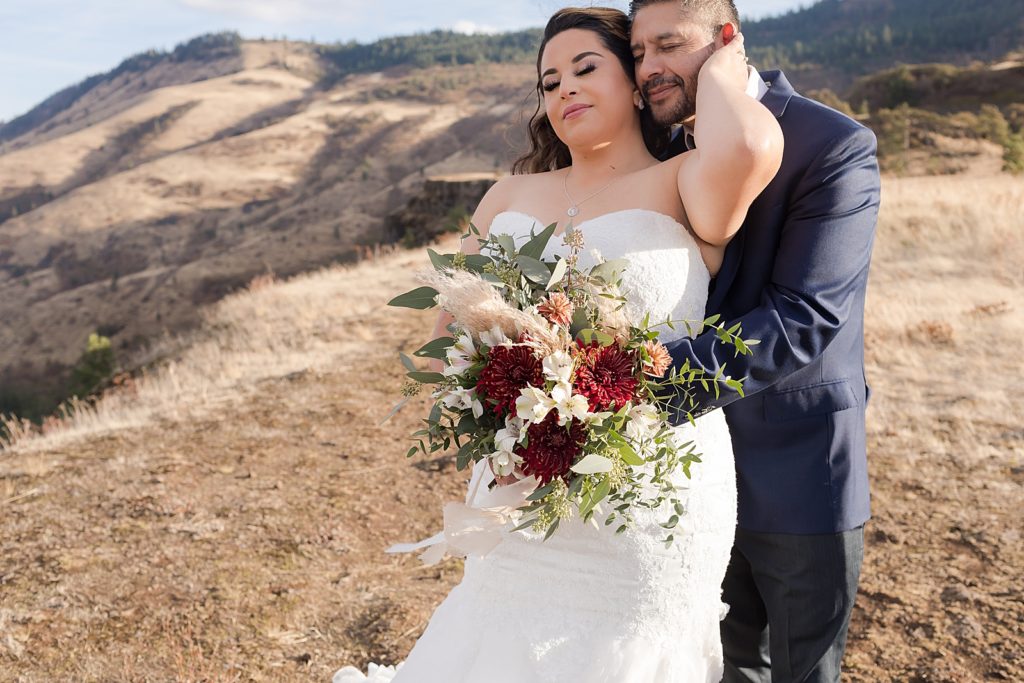 Groom holding Bride from behind holding bouquet on top of mountain