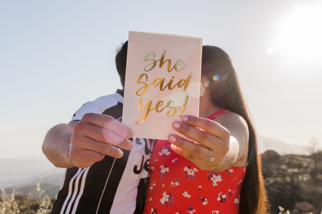 Couple holding up card saying "She Said Yes!" and kissing behind it Santa Barbara Proposal Photography captured by Oregon Engagement Photographer Simply Wandering Photography 