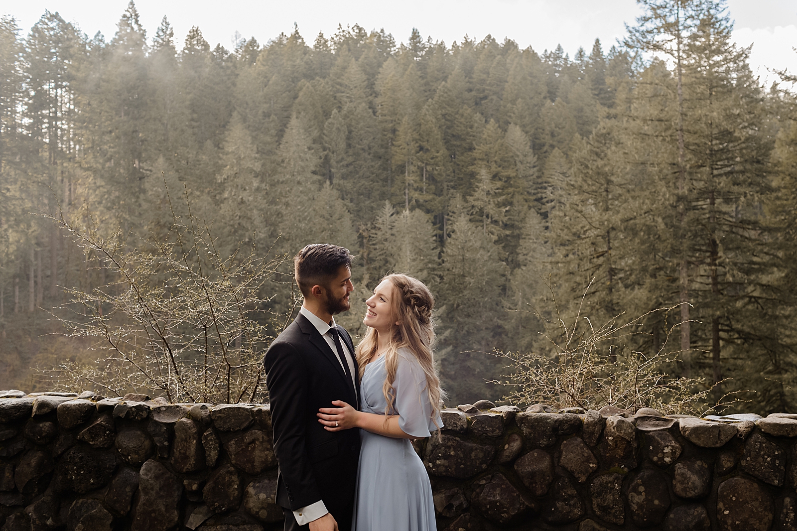 Couple holding each other with trees behind them by a stonewall Silver Falls State Park Oregon Adventure Photography captured by Simply Wandering Photography 